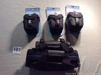 Three black digital camera and camcorder pouches displayed, showing front view attached on store card backing and a larger black padded strap or bag beneath.