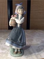 Full front view of the figurine showing the girl standing holding a barrel basket with pink flowers, on a pale tablecloth and neutral background.