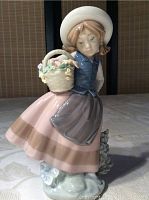Front and side view of the Lladro figurine showing the girl's face, bonnet, basket of flowers, blue vest, and long pink skirt with apron.