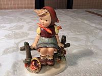 Front view of the Goebel Hummel figurine showing the child sitting on a rustic fence wearing a red headscarf and blue skirt with a basket on the base.