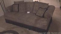 Front view of taupe upholstered sofa with loose pillows, showing fringe skirt