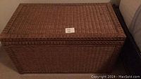 Front view of a woven rattan rectangular chest with a flat lid and brass handles on each side, placed against a wall near a sofa.
