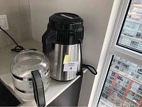Water distiller stainless steel unit next to glass collection jug on a kitchen counter with city view window in background