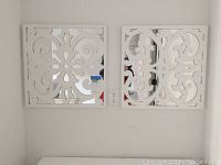 Two white decorative square wall mirrors, mounted on a light wall, showing detailed symmetrical cut-out floral and scroll design.