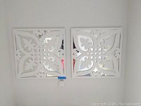 Two white decorative wall mirrors mounted on white wall, showing front view of symmetrical cut-out patterns.