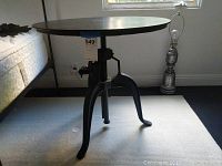 Side view of the round metal table showing the metal tripod base with curved legs, central adjustable crank, and flat round tabletop.