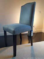 Side view of the blue-gray cloth covered side chair showing fabric texture, high backrest, and dark wooden legs on a reflective floor.