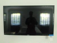 Front view of Samsung 40-inch flat screen TV mounted on white wall with screen off, reflecting light and window behind.