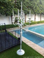 White metal outdoor candelabra standing on concrete poolside with five taper candles in holders. Shows some wear and scratches.