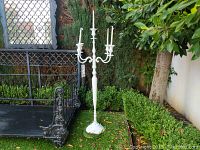 White metal candelabra standing outdoors next to a black wrought iron bench, surrounded by greenery.