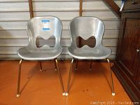 Front view of two vintage metal yard chairs side by side on an orange floor with metal garage door background.
