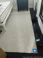 Photo showing large white area rug under a bed frame, visible wear and slightly ribbed texture.