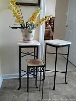 Photo showing three metal plant stands of varying sizes, two with square white tile tops, one smaller with decorative sunflower tile top. Pot with faux flowers placed on one tall stand.