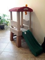 Front-left angled view showing red roof, beige and brown supports, green slide extending to floor, open sides, and upper play platform.