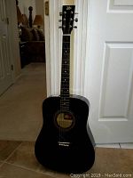 Full front view of the black Rogue acoustic guitar standing upright against a door, shows dreadnought body and six strings.