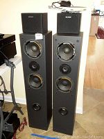 Photo showing two small Sony SS-SR9 cube speakers placed on top of two larger floor-standing Sony SS-AV44 speakers against wall. The large speakers have visible damage and missing front covers.