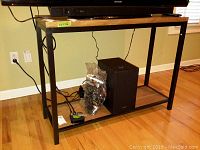 Sofa table with black metal frame and wooden top, lower shelf visible with items placed on it, set against a beige wall with TV mounted above.