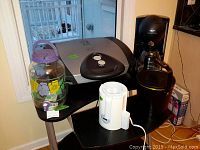 Photo of a black and silver George Foreman Lean Machine grill with two white control knobs, a Krups black drip coffee maker with glass pot, white Munchkin bottle warmer, black copper-tone Presto deep fryer, and large glass juice jug with purple lid and floral design on table.