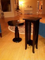 Photo of two black wood tables on wood floor; one taller square table with open storage shelf and a shorter round table with pedestal base.