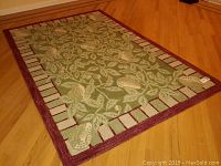 Full view of rectangular wool area rug showing green leaf pattern with bird motifs and maroon bordered edge on hardwood floor.