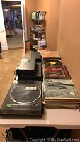 Turntable system and stacks of vinyl LP records on a folding table in a room with tile flooring.