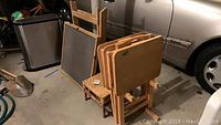 Photo of four folded wood TV trays placed on a wooden stand and a small wooden stool, with a chalkboard behind (not included). The items are in a garage setting next to a car.