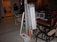 Two ceiling shop lights and packages of fluorescent bulbs stacked on floor in basement with some chairs and books