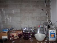 Overview of glassware including purple amethyst pitcher and goblets on left, various boxed items, plastic plates and other serveware on right.