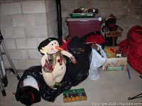 Three plush seasonal dolls (snowman, bear, moose), box of Christmas ornaments on top of a plastic storage bin, wreath, and other seasonal decorations in basement storage area.