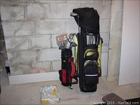 Photo showing two golf club sets side by side in bags, along with a box of white golf balls on the floor, set against a concrete block wall.