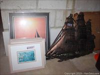 Photo showing one large metal ship wall art sculpture with five framed artworks stacked against a wall.