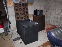 Photo showing the two large black Aiwa speakers positioned on the floor in a basement environment with concrete and cinder block walls. Nearby wooden Aiwa stereo system also visible.
