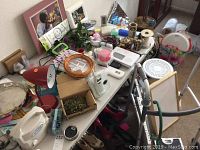 Overview of various household and bathroom items on a table, including decorative frames, containers, artificial flowers, a red desk lamp, white digital scale, and miscellaneous small items.