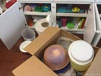 Plastic food storage containers inside a cardboard box and kitchen cabinets, showing multiple lids and bowls in different colors.