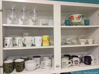 Shelf with multiple coffee cups, mugs, glassware and ceramic bowls including glass mugs with stems, branded mugs, and decorative ceramics.