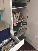 Wide view of open white cabinet showing shelves of cookbooks, cookware, glassware cups, and a drawer with silver plate flatware case.
