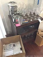 Bar cabinet with crystal glassware displayed on top and shelves; additional accessories around including small glass and stainless container.