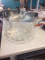 Covered glass cake stand with knob handle lid and floral embossed plate, photographed on countertop to show design and shape
