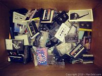 A box filled with over 30 audio cassette tapes, some loose, showing labels and handwritten notes.