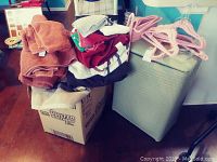 Photo of folded linens, towels, washcloths atop boxes and a woven-style beige laundry hamper with pink plastic hangers on top.