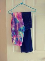 Dark blue denim jeans folded with colorful tie-dye leggings on a hanger against a white door