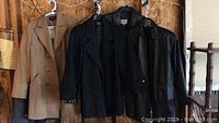 Five women's medium-sized jackets on hangers against wooden wall: camel double-breasted wool jacket, black wool jacket, and three black leather jackets including one knee-length. Visual confirmation of jacket styles and colors.
