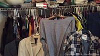 Photo of assorted women’s clothing hanging on wire shelving, including a beige suit jacket, gray top, and other tops and slacks