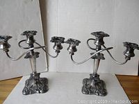 Pair of vintage silver plated three-arm candelabras with decorative floral and vine designs on base and arms, positioned side by side against a plain backdrop.
