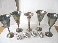 Four silver-plated wine cups, four fleur de lis napkin holders, and one bud vase grouped on a wooden surface, showing the overall set with visible tarnish.