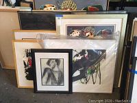 Five framed artworks by Jenik Cook stacked, front shows pencil sketch portrait framed in black and a large abstract painting wrapped in plastic