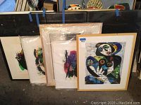 Five framed artworks leaning against a wall, one unwrapped and visible cubist-style piece on right