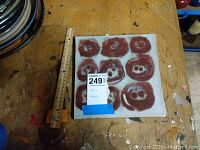 Front side of a square ceramic tile on wooden table. Shows tile with nine red circular abstract faces on gray background and a ruler next to it for scale.