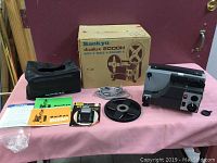 Complete lot displayed on pink cloth: Sankyo Dualux 2000H projector, original box, user guide, quick splice tool, film reel, power cord, and carrying case.