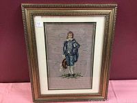 Full view of the vintage Blueboy cross stitch in its gold-toned ornate frame against a burgundy wall.